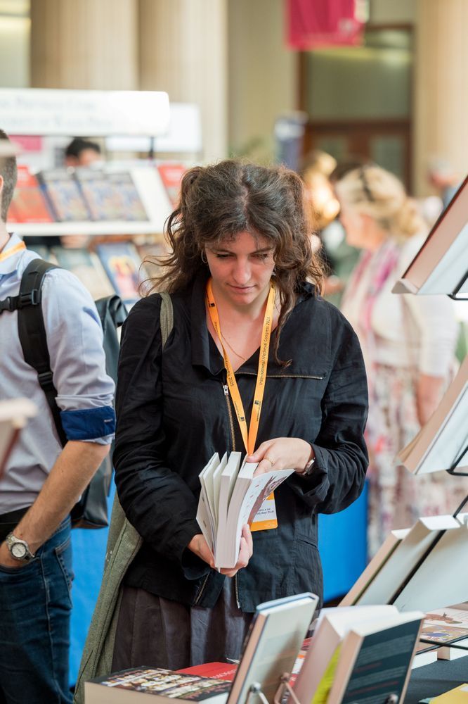 A delegate at the IMC Bookfair, wearing an orange lanyard holding a book and looking through the pages.
