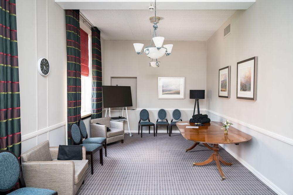 One of the Health and Wellbeing rooms in University House. There are comfy chairs and a table. There is also a tv screen a lamp, a clock and pictures on the walls. 