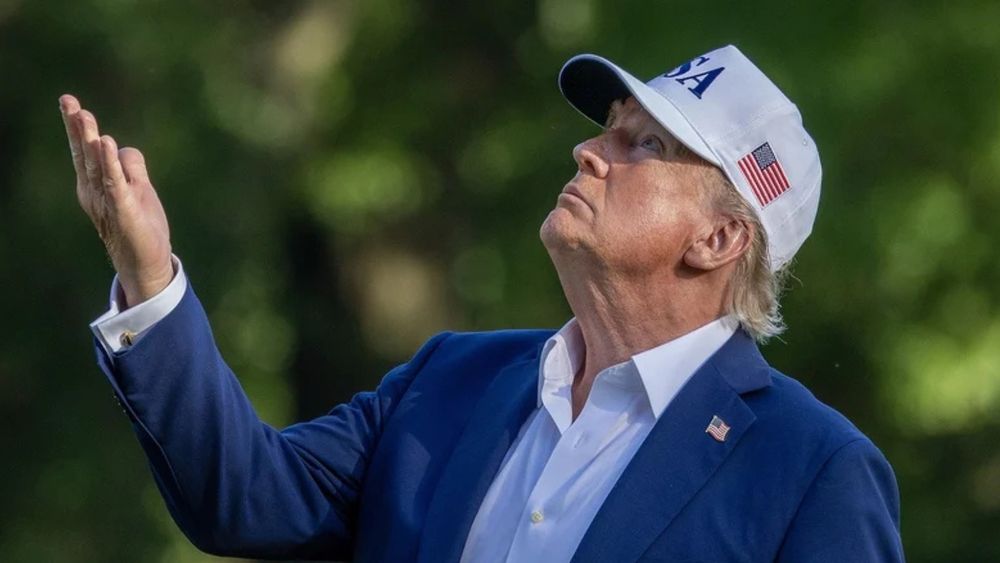 US president Donald Trump. He is looking upwards and has his hand raised in the air. He is wearing a white shirt and blue blazer. He is wearing a white baseball cap that has the American flag on one side and USA emblazoned on the front. There is also an American flag pin on his blazer. 