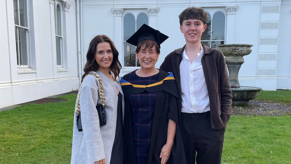 UL graduate Catherine Caball. She is wearing graduation robes and a graduation hat. She is smiling broadly and standing outside a large white 18th century building with a fountain and large white sash windows. She is flanked by her daughter and son who are also smiling broadly.