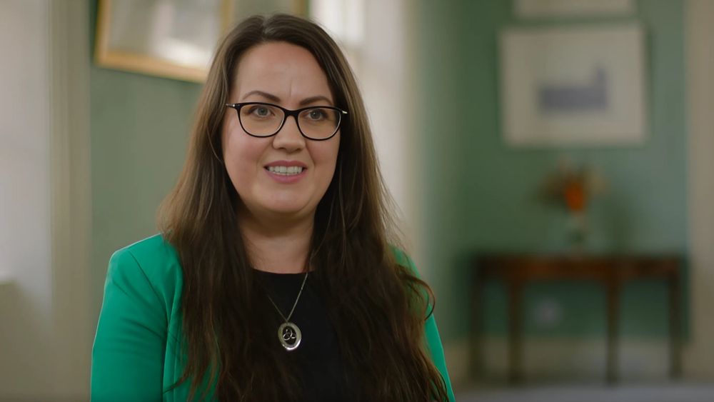 Dr Karol Mullaney-Dignam, Associate Professor in the School of History and Geography. She is seated in a room with mint green walls. She has long brown hair and is wearing glasses. She is wearing a black top, green blazer and silver necklace. 