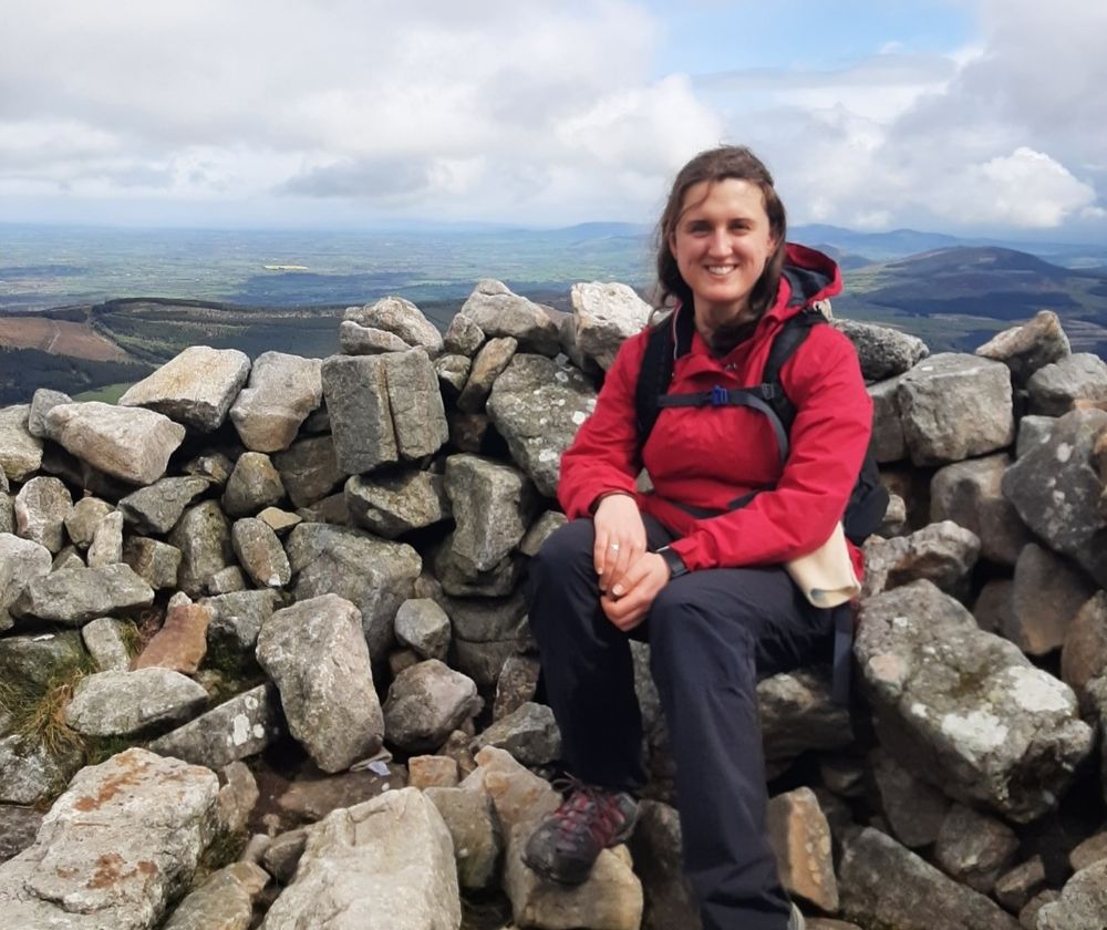 Photo of woman in red jacket sitting on stone wall in mountain setting