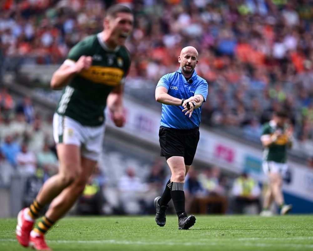 An action picture of referee Brendan Cawley in a blue jersey pointing in the middle of a GAA playing pitch