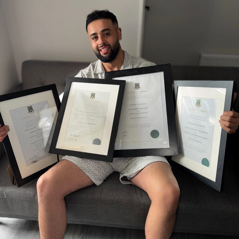 Man smiling for photo in indoor setting holding four framed certificates