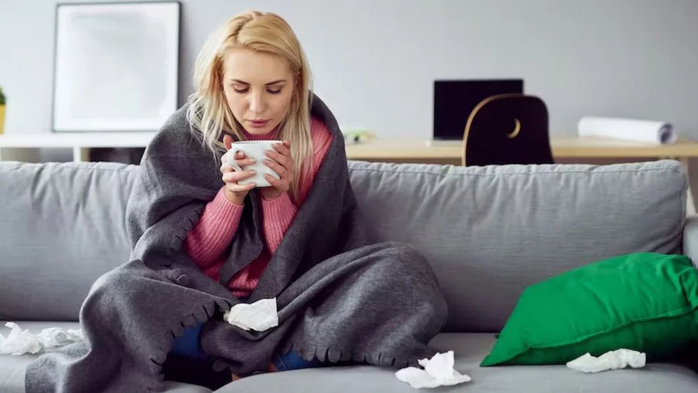 A woman sitting on a grey couch. She is wearing a pink jumper and has a grey blanket wrapped around her. Her hands are wrapped around a white mug. She has blonde hair. There's a green cushion on the couch and a desk with a laptop and chair in the background. 