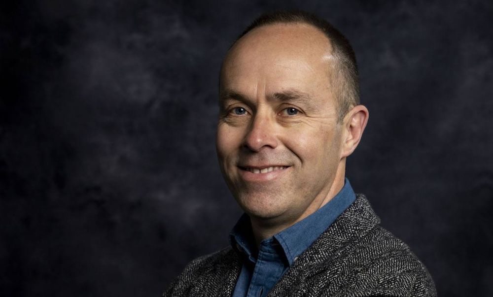 UL lecturer Dr John Garvey. He is wearing a grey tweed blazer and a blue shirt. He is standing in front of a dark grey backdrop. 
