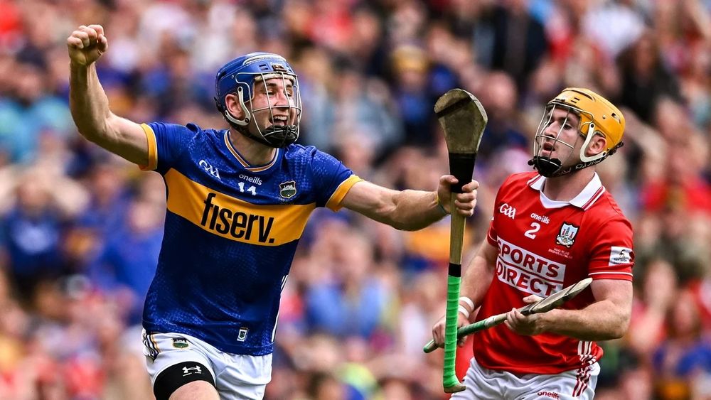 A Tipperary player in a blue and yellow jersey celebrating as he runs past a Cork player in a red and white jersey
