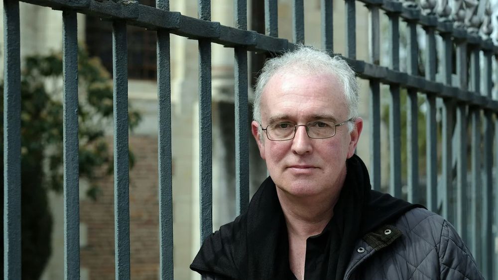 Professor of Creative Writing at UL and best-selling novelist, Joseph O'Connor, standing beside an iron railing. He is wearing a black jacket and glasses. He has grey hair. 
