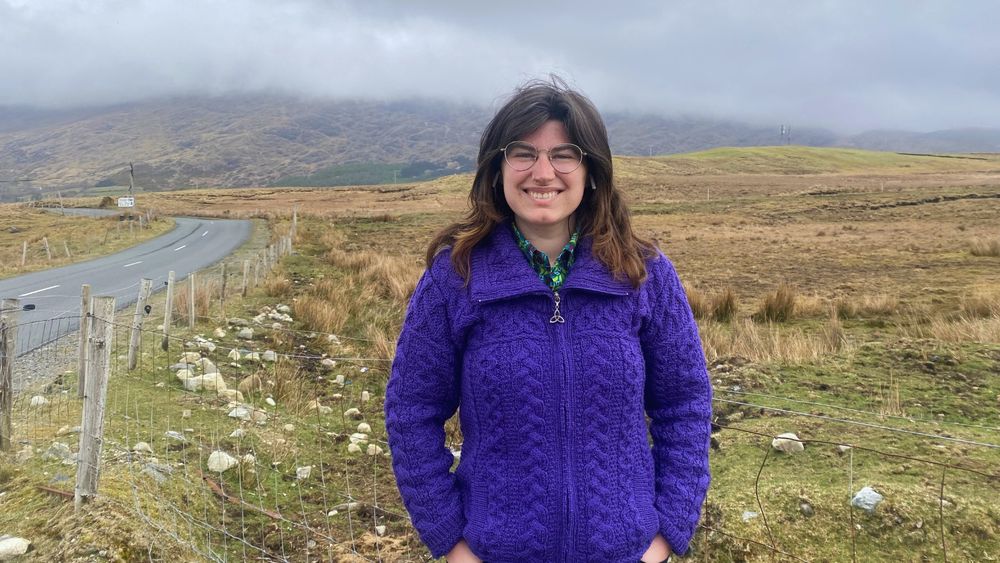 Abigail Hickman, a graduate of the MA in English at UL, stands in front of a field in the west of Ireland. She is wearing a purple cardigan. 