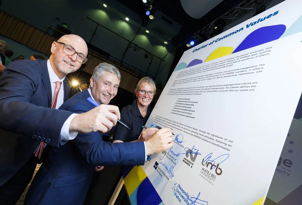Photograph of three people in smart work attire stood next to a large information board. They are signing their names next to the logos for each of their universities as they sign up to a charter