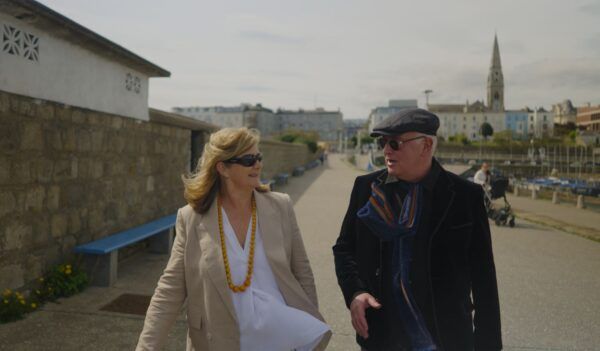 Professor of Creative Writing and best-selling novelist Joseph O'Connor walking with Madeleine Keane, literary editor of the Sunday Independent. They are in conversation as they walk along Dún Laoghaire pier. There are large buildings and a church steeple in the background. Joseph is wearing a black blazer, a tweed flat cap and navy striped scarf. Madeleine is wearing a white shirt and beige blazer. She is also wearing a large necklace with yellow beads. Both are wearing sunglasses.