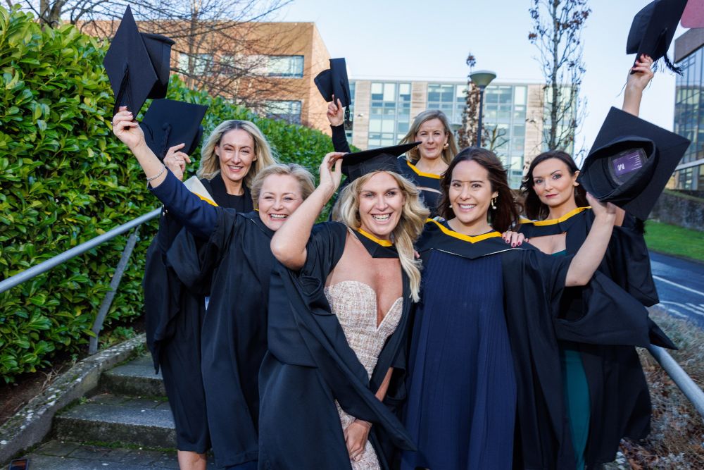 A group of six graduates in black and gold gowns over their dresses all raising their caps in the air in celebration while smiling at the camera
