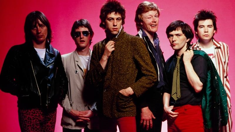 Irish rock group The Boomtown Rats. There are six members of the band standing in front of a pink background. Lead singer Bob Geldof is in the middle dressed in a brown tweed jacket and red trousers. The other band members are standing on either side of him with serious faces while one band member winks. 