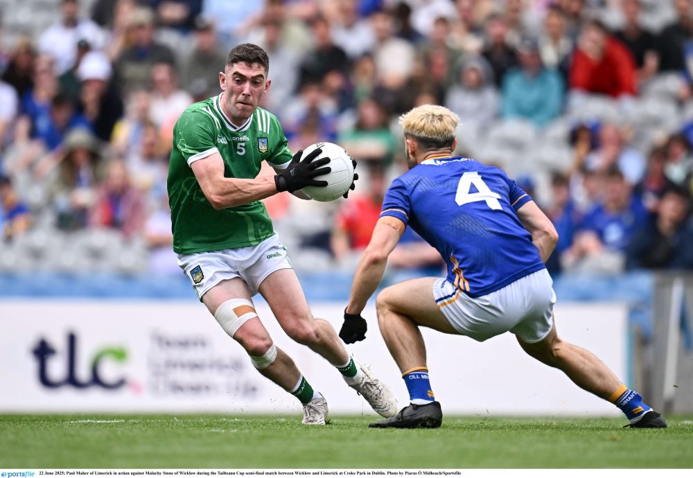 Photograph of two footballers, one in a green jersey and white shorts, holding the ball and running with it. A player in a blue jersey and white shorts is attempting to tackle him