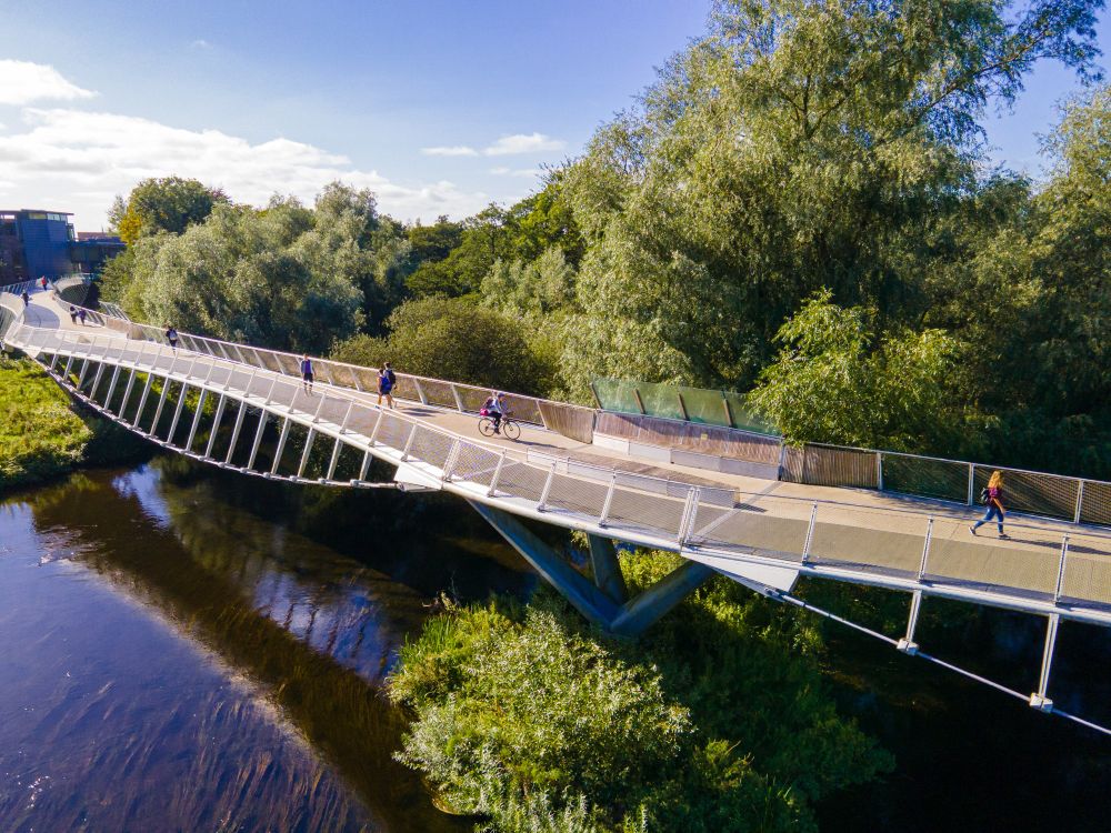 The living bridge on the UL campus. The River Shannon flows under the bridge and there are trees full of green leaves in the background. There are people walking across the bridge and a person cycling. The sun is shining and the sky is blue. 