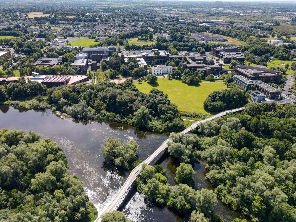An aerial photograph of the University of Limerick campus showing a large river spanned by a curved pedestrian bridge. The river is lined by dense trees. There are several lawns and large academic buildings nestled between the trees