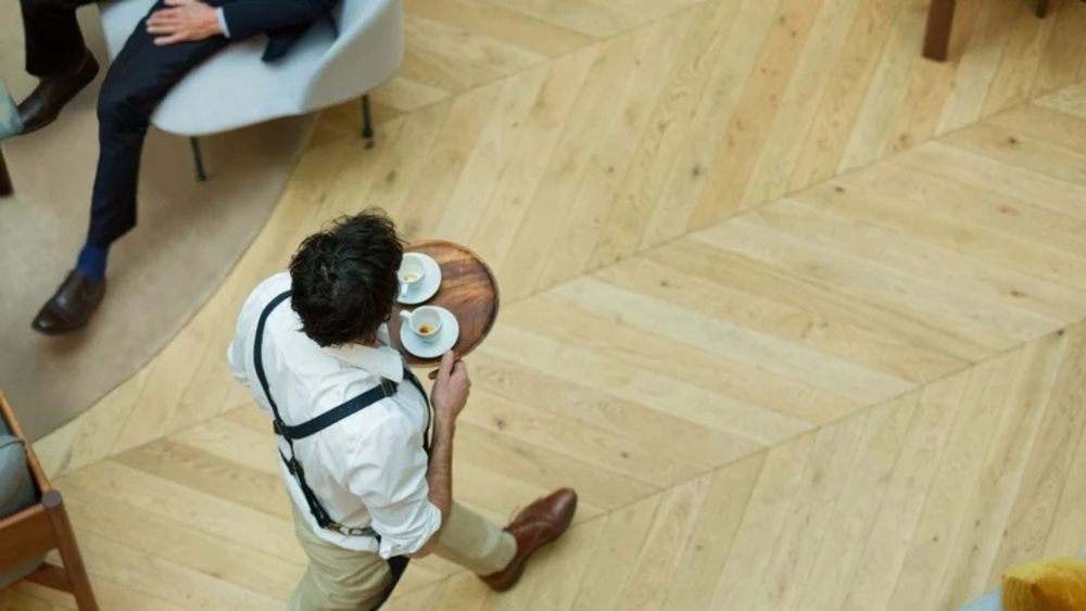 An aerial view of a waiter dressed in a white shirt, cream trousers, navy apron and brown shoes. He is walking across a wooden floor and holding a wooden tray with two cups of coffee. 