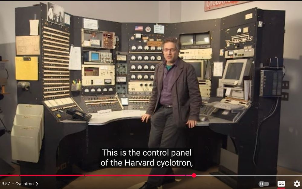Peter Galiston, faculty director of the Harvard Collection of Historical Scientific Instruments standing in front of the control panel of the Harvard Cyclotron.  The control panel includes knobs and switches from the 50's, oscilloscopes of the 60's and 70's, and two PCs from the late 1990s. 