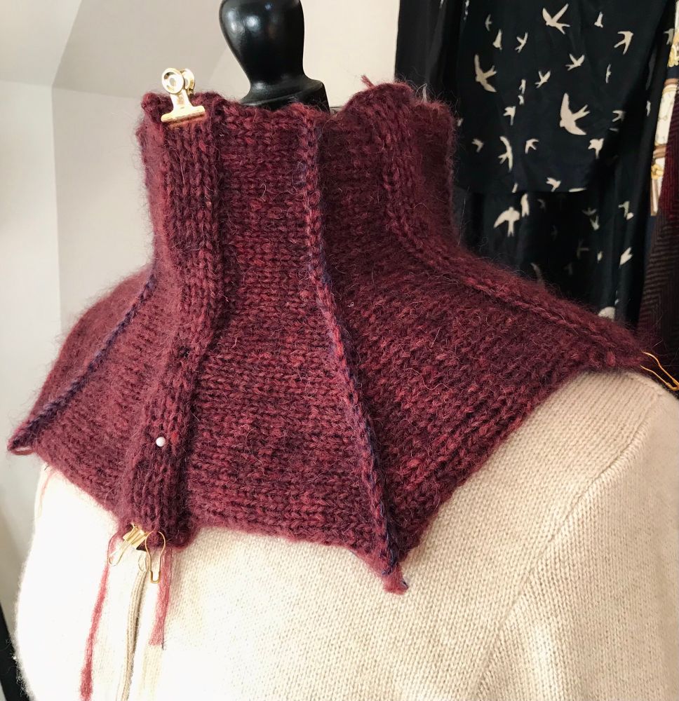 Close up of a work in progress of a handknitted collar in burgundy with a subtle contrasting blue colour. It is shown on a mannequin wearing a beige cardigan in front of a wall with hanging shawls. 