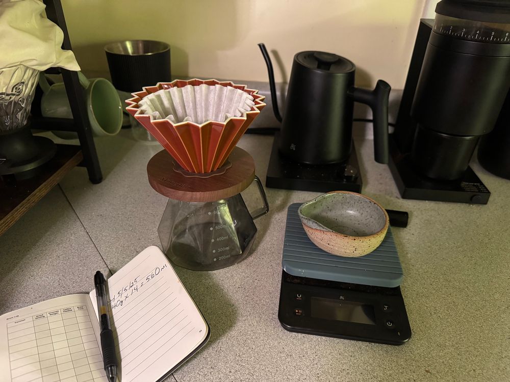 Origami ceramic coffee dripper on carafe. Black gooseneck kettle and grinder, hand made ceramic dosing cup on scale, small notebook and pen to the left.