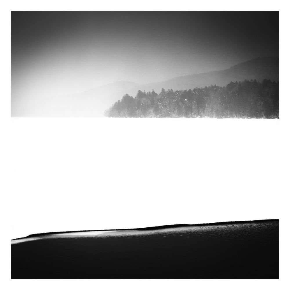 A serene black and white landscape photo taken at Lake Akan in Japan. Dark black water in the foreground contrasts with ice on the lake surface. Trees and a mountainside appear in the distance hazy behind a snow shower.