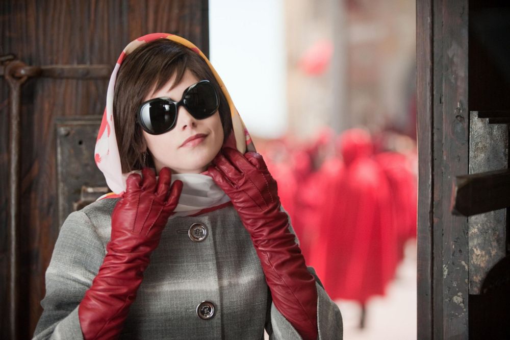 Movie still of Alice Cullen (played by Ashley Greene) prepares to remove her pink, yellow, and white headscarf to greet the Voturi. She is wearing giant black sunglasses, long red leather gloves, and a grey coat. In the background, people in red cloaks mull about a festival.