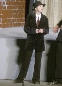 Picture of a young person in their late teens or early 20s wearing a suit, tie, and a fedora on a stage.