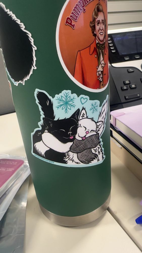 Sticker of a black cat and a white cat, both wearing scarves and snuggled up together with a little blep on each of them. Yes they look like Ed and Stede to me. Above them is a sticker of Pumpkin Spice Stede made by CitrusSyndicate. All stickers are on a tea thermos on my work desk.