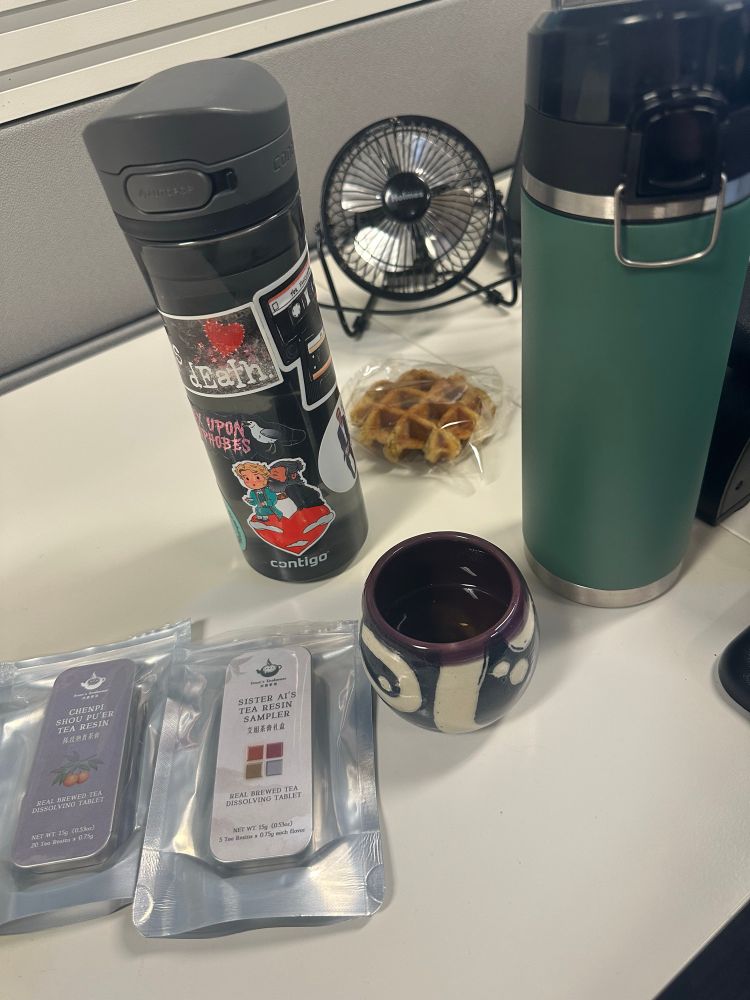 Picture of a desk with 2 packets of tea resins, a hand made ceramic tea cup, thermos, water bottle with OFMD stickers, a liege waffle and a little desk fan