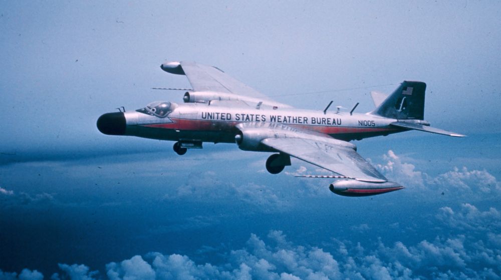 colour photo of a B-57 variant in flight, banking to the camera high in a cloudy blue sky, with the undercarriage down. the aircraft is silver, with orange lightning bolts down the fuselage and wingtip pods. “UNITED STATES WEATHER BUREAU” is written in large lettering along the fuselage. there is also a black “dog nose”, and striped probes on each wing 
