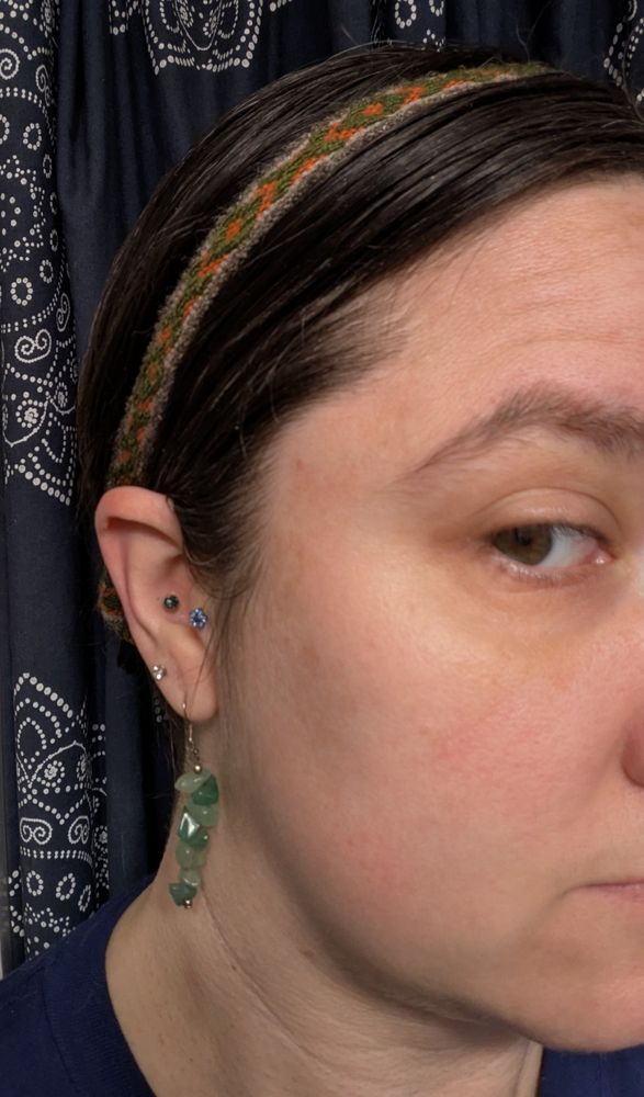 A white woman with brown hair is standing in front of a navy blue curtain with white mandalas.  She has a handwoven band tied over her hair.  It is mostly orange with elongated X and O motifs in olive green.  There are four earrings in her ear, one of which is a stack of polished green aventurine about an inch long.