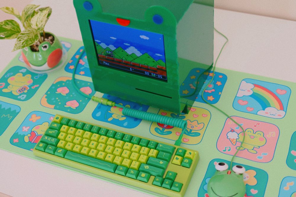 frog shaped computer and green keyboard with frog themed icons, on top of a colorful deskmat with cute frog drawings. the frog computer screen shows a level from a keroppi super nintendo game. a frog planter and frog computer mouse are also on the desk.