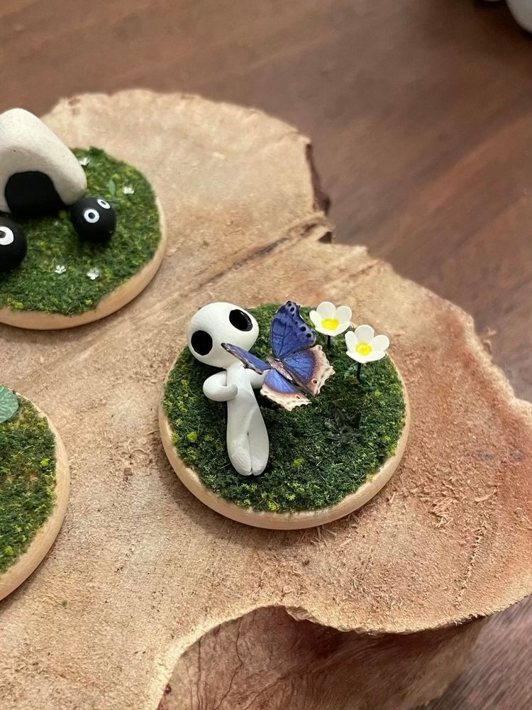 a tiny diorama with fake grass, tiny flowers, a butterfly, and a kodama. in the background is another tiny diorama with soot sprites and onigiri