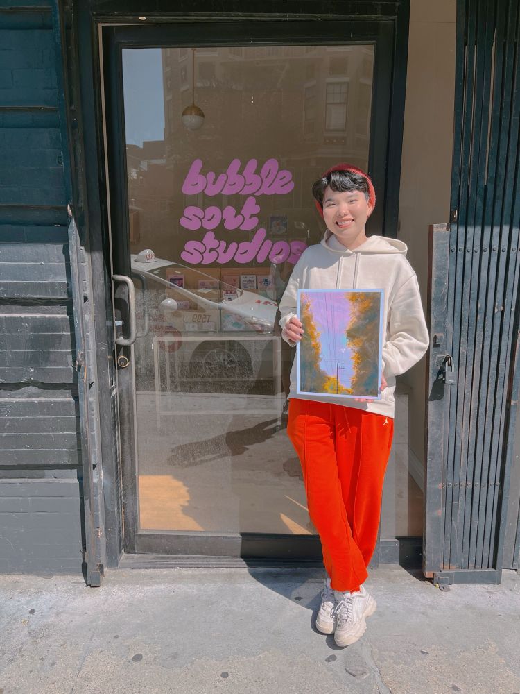 asian woman with short hair, red hat, white hoodie, and red pants holding a print of trees and a power line set against a colorful sunset, standing in front of a glass door with pink bubble letters spelling out “bubblesort studios”