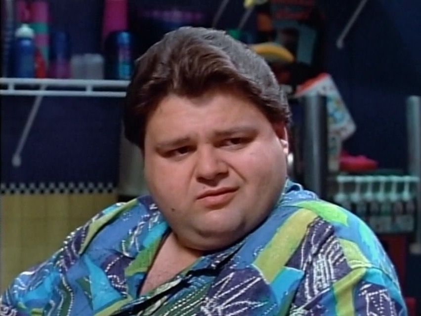 Ernie from the power rangers tv show wears a fun Hawaiian style shirt and looks around in snarkiness at all the kids at his juice bar and martial arts studio.