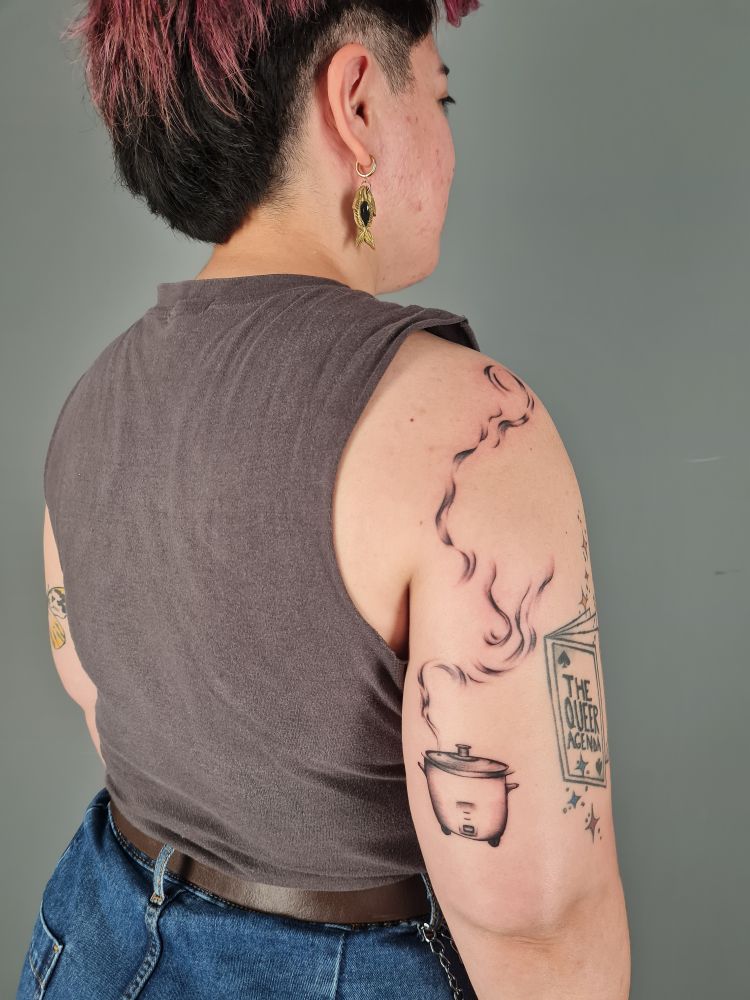 Photo taken of the back of Sarah's right arm. There is a tattoo of a rice cooker above their elbow with steam moving upwards to their shoulder, where it swirls. To the right of the tattoo is an existing tattoo of a zine with "the queer agenda" on the front and different coloured stars above and below it. Sarah is wearing a fish earring on their right ear.