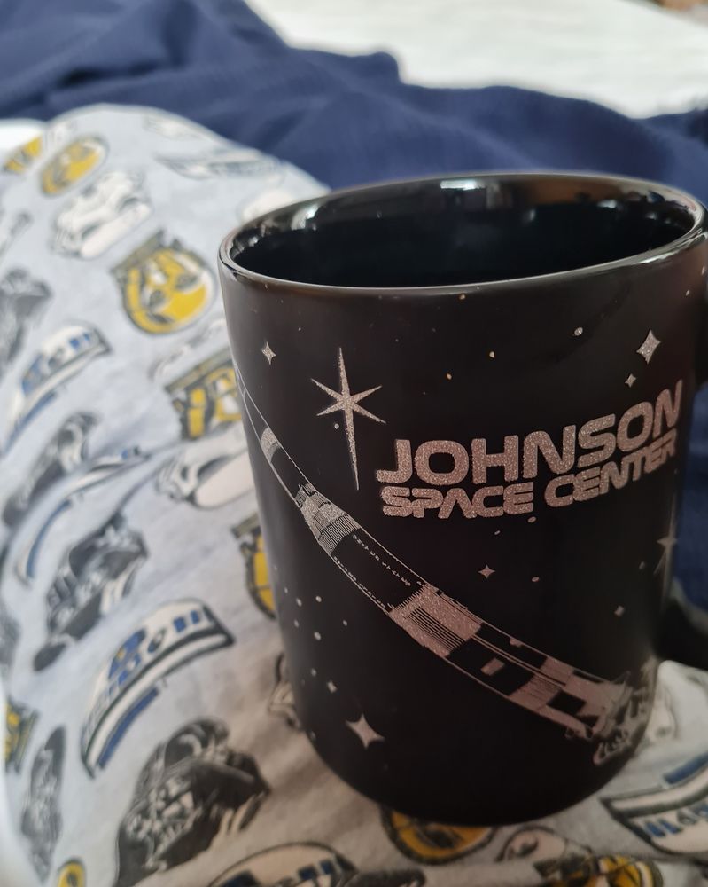Photo of a black mug with a space shuttle and "Johnson Space Centre" in silver. Sarah is wearing pajama bottoms with a print of C3PO, R2D2 and Darth Vader. The mug is resting on top of the leg.