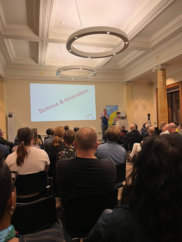Room with people sitting down listening to Sarah speak, who is behind a podium with a BSL interpreter standing next to them. Behind them is a slide projected which says 'science and innovation'.