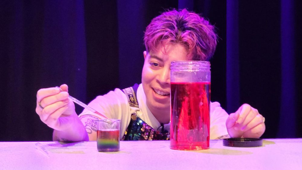 Sarah squatted down to pipette red liquid into a small beaker on a table. The beaker has red, orange, yellow, green, blue and purple layers of liquid. Partially blocking Sarah's face is a container with red liquid.