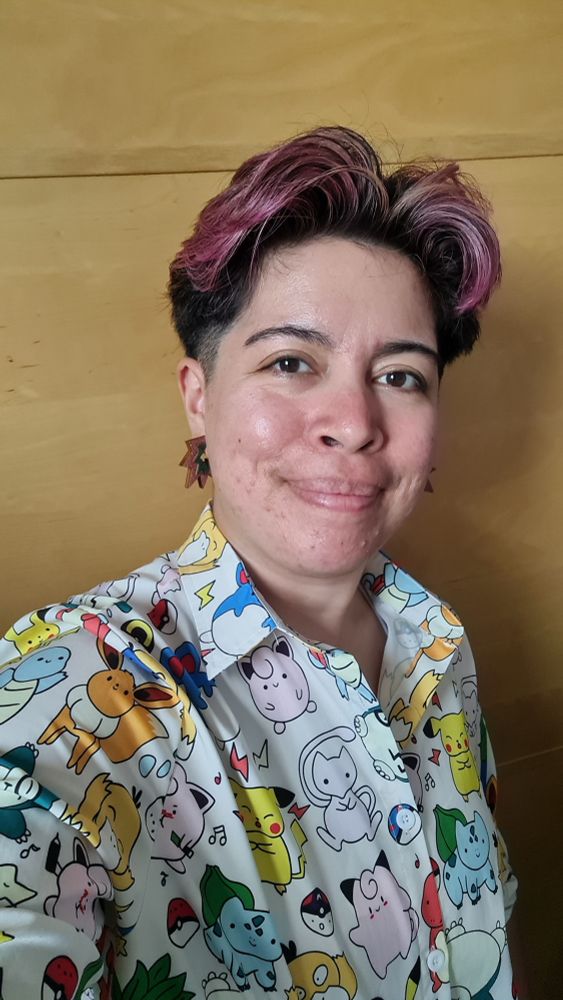 Sarah is wearing hoop earrings which have a zigzag design to look like a small explosion, with pink on the outside and green on the inside. They are also wearing a white button up with a print of different pokemon. Sarah also has pink short hair.