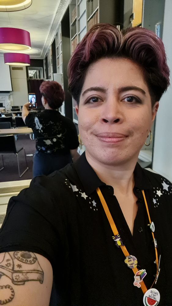 Sarah taking a selfie of themselves with a mirror behind them. They are wearing a black button up shirt with three silver stars and dots on each side, around the collarbone. The mirror shows the back of the shirt which has embroidery of a space shuttle, planets and stars. Sarah is wearing a lanyard with multiple pins: a frog with non binary flag colours; a she they pronoun pin with non colours; a megaphone with the progress flag coming out of it; a badge which says "science is not neutral" with a watermelon illustration underneath; a cell; and two other pins turned away so you can't see the design. Sarah is also wearing gold metal earrings bent into a curved design.