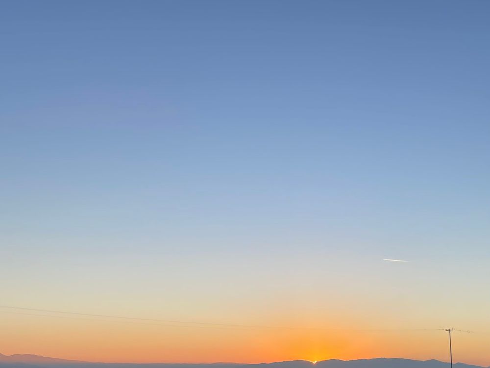 Image of mostly blue sky at dusk with just a pop of bright orange sunlight peeking just above a distant row of purple mountains on the horizon. 
The silhouette of a power line is seen where the burnt orange of the horizon morphs into ombré hues of orange -yellow-green-blue.