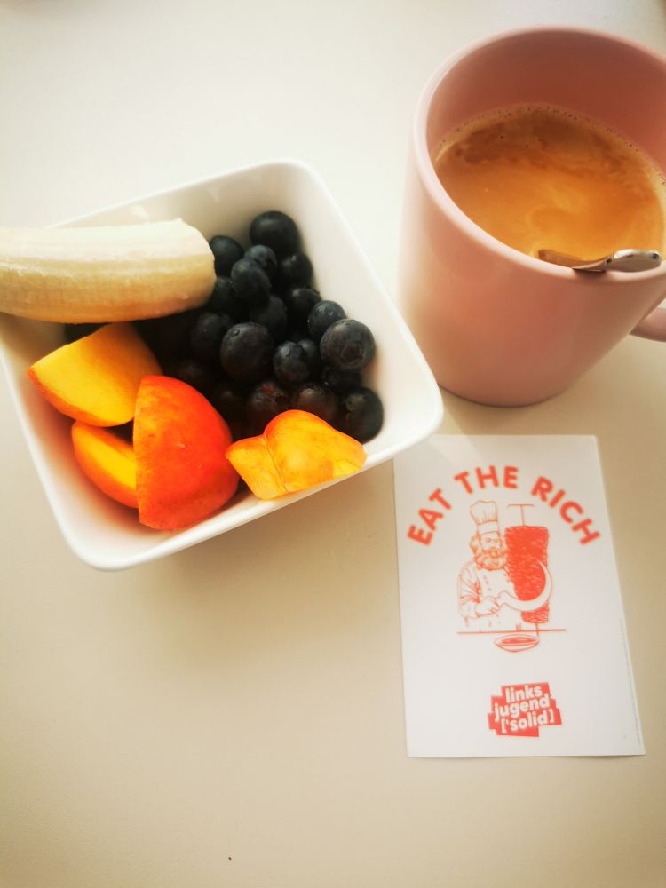 Schale mit Blaubeeren, Nektarinenstücken und Bananenstücken, eine Tasse Kaffee und ein Sticker mit dem Aufdruck Eat the Rich