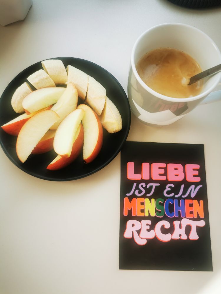 Ein Tellerchen mit Bananenstücken und Apfelstücken eine Tasse Kaffee und ein Sticker mit dem Text Liebe ist ein Menschenrecht 