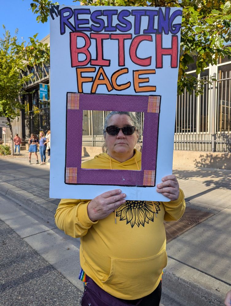 Two photographs side by side with a woman holding a sign that says "resisting b**** face." There is a cutout square under the headline where she can put her face in a frame.