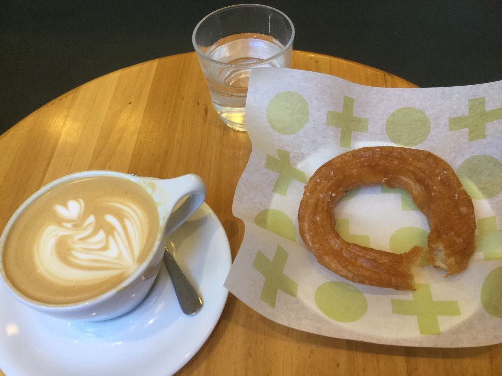 A cappuccino in the lower left corner, a cruller with a bite taken out on the right, a small glass of sparkling water at the top.