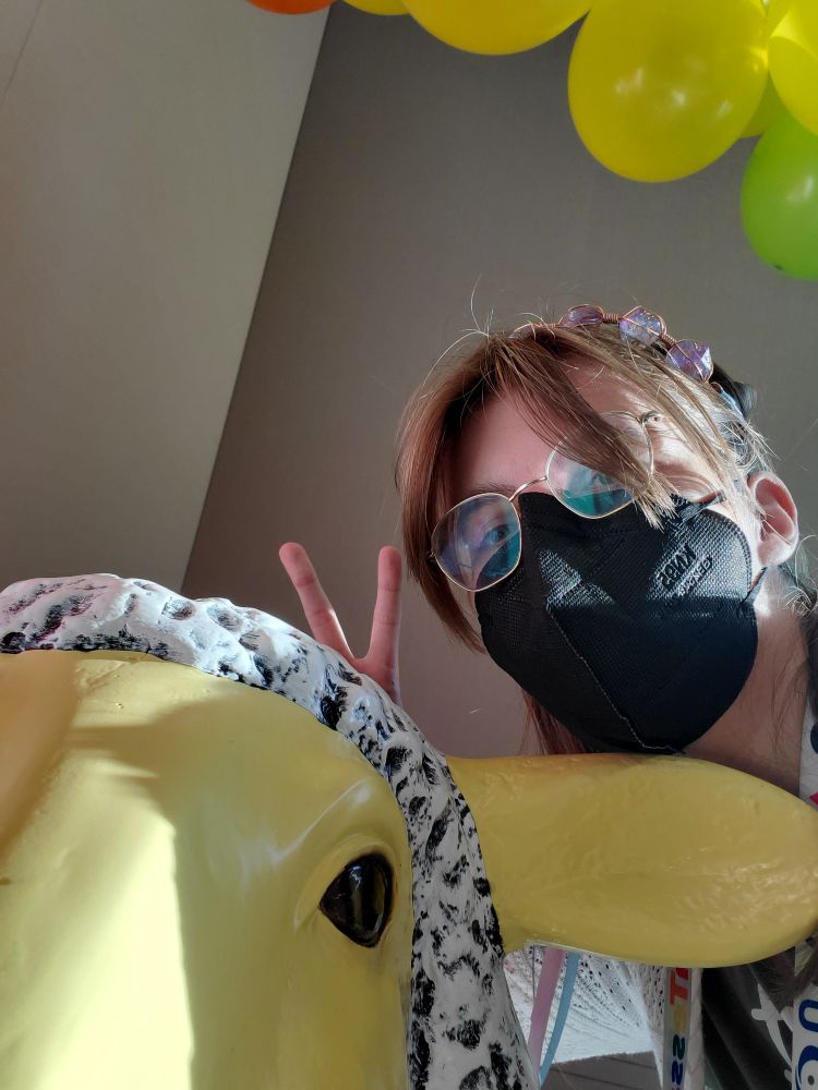 A white person with glasses, a face mask, and a dice crown gives bunny ears to a large sheep figurine named Lady Ba Ba.