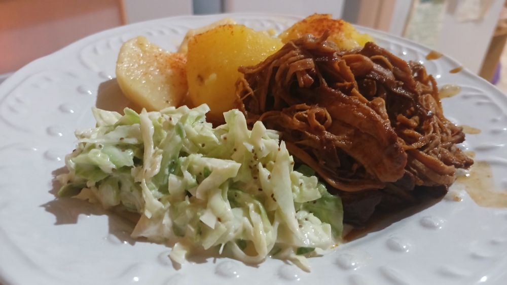 A white plate holds green coleslaw with a creamy dressing and celery seed,  furls of saucy bbq pork, and slow roasted tender potatoes. 