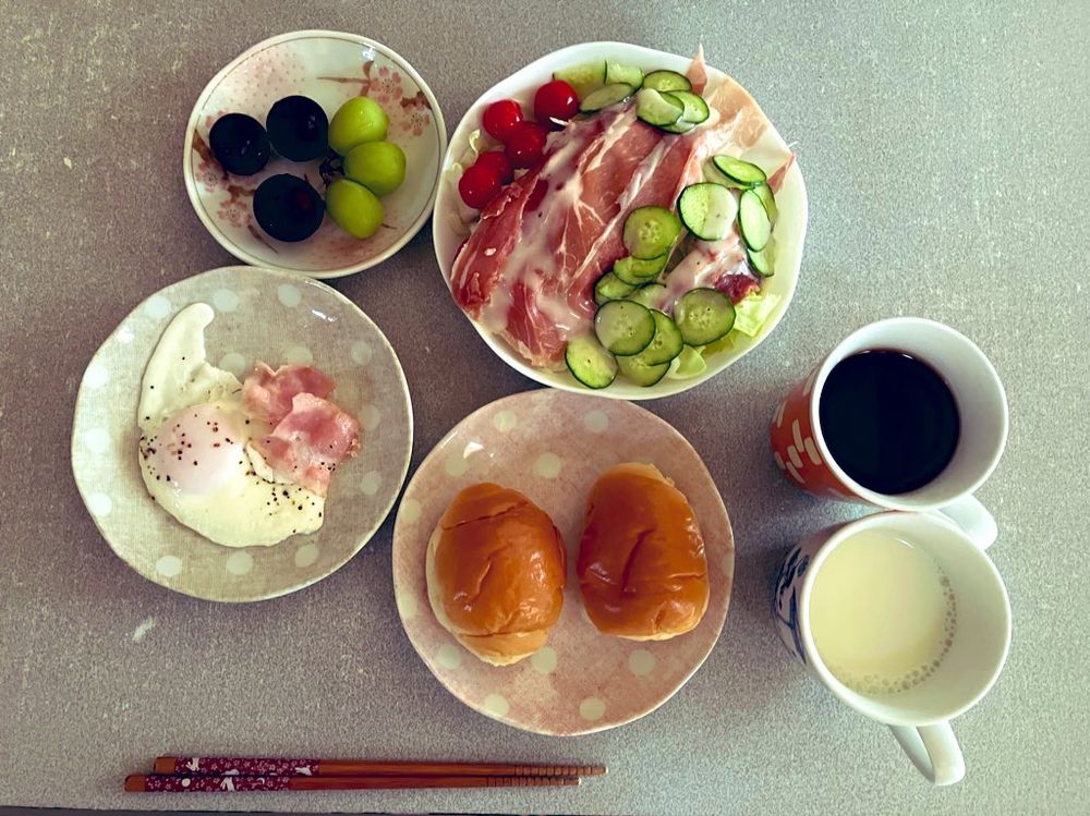 右側に牛乳の入ったマグカップと紅茶の入ったマグカップ。手前にお箸。真ん中にバターロールパン２個。その隣に目玉焼きにベーコン。目玉焼きの皿の上にバイオチェリー３個とシャインマスカット３個。真ん中の大きめな皿にサラダ。キャベツの千切りとハモンセラーノハム、ぷちぷよ、きゅうり、シーザードレッシング。