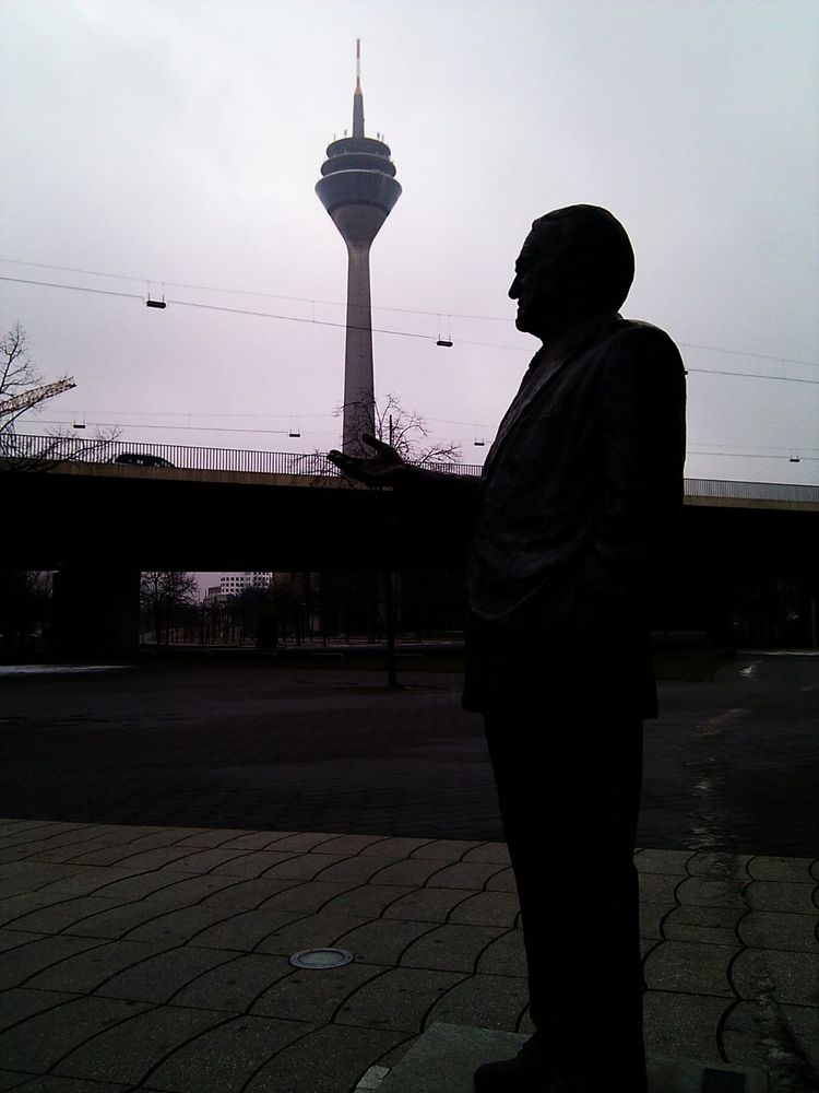 Gegen das Sonnenlicht fotografiert, wodurch man fast nur die Konturen erkennt. Auf der rechten Seite das Denkmal von Johannes Rau, mittig über der Rheinkniebrücke der Rheinturm, der durch die bewusst gewählte Perspektive auf der ausgestreckten Hand von Johannes Rau zu stehen scheint.